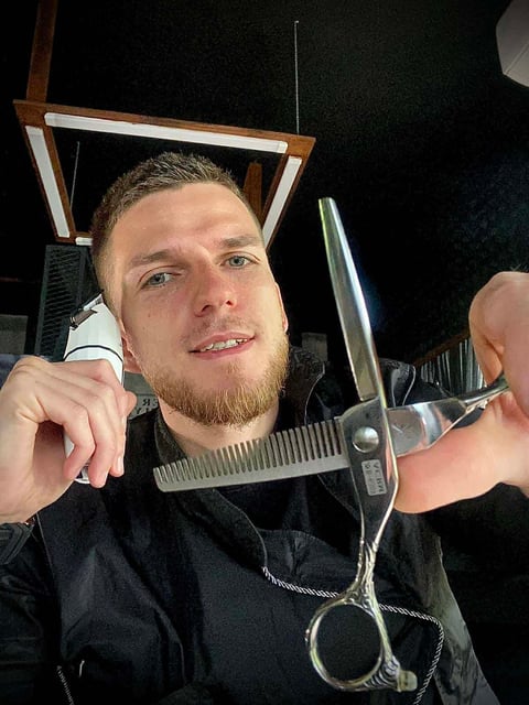 Man holding barber scissors and comb in front of a mirror, smiling at camera in salon setting